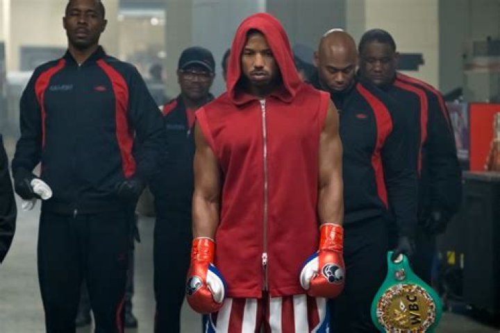 Adonis Creed (Michael B. Jordan), in red sweatpants, a gray tank top, and white boxing gloves, crouches in the corner of a gym boxing ring and looks grimly determined in Creed III