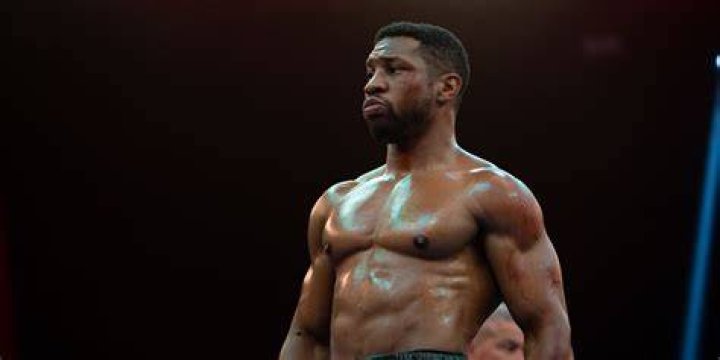Damian (Jonathan Majors), a boxer in a grey sweatshirt, black sweatpants, and electric blue boxing gloves, leans down out of a boxing ring in a gym to bump fists with Adonis Creed (Michael B. Jordan) in Creed III